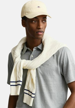 Mann mit beiger Mütze, hellgrauem Poloshirt und cremefarbenem Strickpullover mit Zopfmuster, der über die Schultern gelegt ist, mit dunkelblauen Streifen an den Ärmeln.