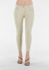 Jeans beige aderenti con vita alta, due tasche frontali e dettagli in metallo argentato. Abbinati a sandali neri con tacco alto e cinghie.
