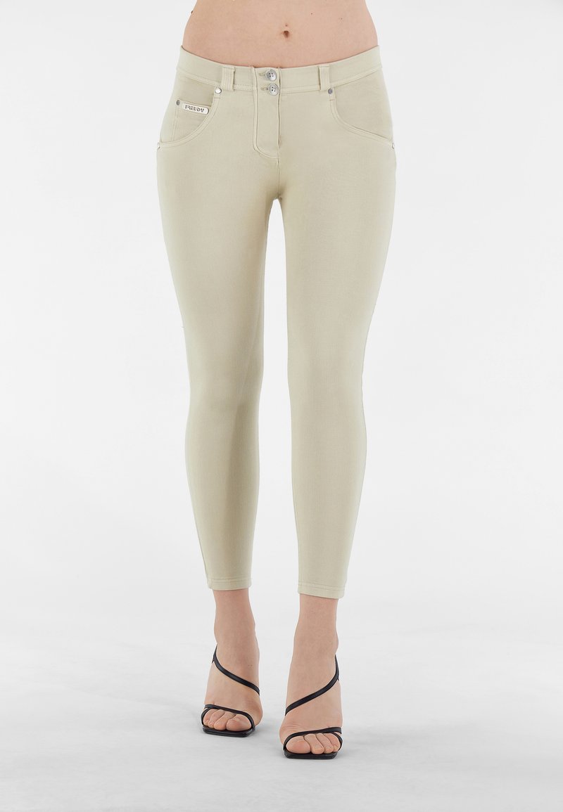 Jeans beige aderenti con vita alta, due tasche frontali e dettagli in metallo argentato. Abbinati a sandali neri con tacco alto e cinghie.
