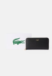 Portafoglio in pelle nera con una finitura texturizzata, caratterizzato da un logo dorato e da una chiusura con zip. Accompagnato da una scatola bianca con un logo di coccodrillo verde.