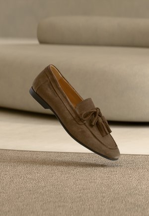 Hnedý semišový mokasín s strapcami vznášajúci sa nad texturovaným béžovým kobercom a pozadím zodpovedajúcej béžovej pohovky.