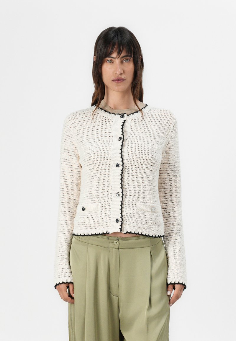 Cardigan a maglia color crema con rifiniture nere, dotato di scollatura rotonda, bottoni frontali e due tasche anteriori. Dettagli a punto testurizzato in tutto il capo.