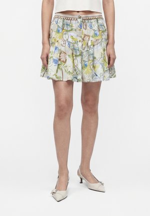 Žena nosí krátku, vzdušnú sukňu s farebným kvetinovým a botanickým vzorom, doplnenú bielymi nízkymi sandálmi so stuhami na päte.