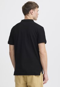 Černé polo tričko s krátkým rukávem vyrobené z bavlny, s klasickým designem, rovný lem a boční rozparky, pohled zezadu.