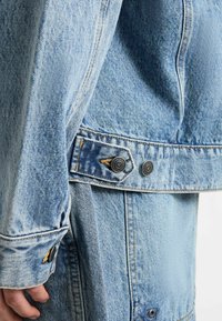 Hellblaues Denim-Jacket mit auffälligen goldenen Nähten, großen Knöpfen und ausgeprägter Textur, das ein klassisches Design mit taillierter Passform präsentiert.