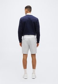 Άνδρας γυρισμένος προς τα πίσω φοράει σκούρο μπλε πουλόβερ, λευκό πουκάμισο με γιακά, ανοιχτό γκρι σορτς, λευκές κάλτσες και λευκά αθλητικά παπούτσια, σε απλό φόντο.