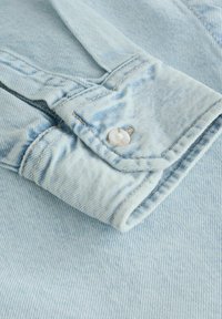 Poignet de chemise en denim bleu clair avec bouton blanc et surpiqûres visibles sur un fond en tissu denim assorti.