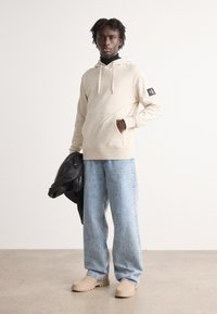 Felpa beige con cordini e tasca frontale, abbinata a jeans larghi azzurri e zoccoli beige. Giacca in pelle nera appoggiata sul braccio.