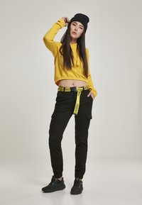 Žuta skraćena dukserica, crne cargo hlače s žutim pojasom "URBAN CLASSICS", crne patike i crna kapa. Elegantan i funkcionalan izgled.