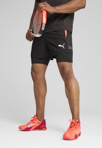 Schwarze Sportshorts mit weißem Logo und roten Akzenten; das Modell hält ein Paddelracket. Helle rote Sportschuhe mit strukturierten Oberflächen und schlankem Design.