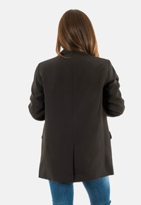 Blazer noir à manches longues, avec fermeture à un bouton et deux poches avant. Le tissu semble lisse avec un léger reflet.