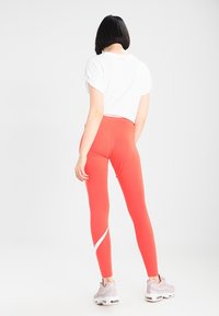 Žena s krátkými černými vlasy, oblečená v bílém tričku, korálových legínách s bílým pruhem a světle růžových teniskách, stojící na prostém bílém pozadí.