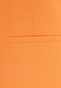 Oranžová látka s texturovaným tkaním. Obsahuje jednu plochou kapsu na pravé straně, která je jemně integrována do designu.