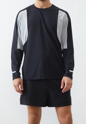 Mann trägt schwarzes, langärmeliges Sportshirt mit weißen Einsätzen an den Ärmeln und schwarze Sportshorts, steht vor weißem Hintergrund.