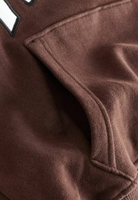 Gros plan d'une poche en tissu marron avec des coutures visibles et un bord plié sur un vêtement à texture douce.
