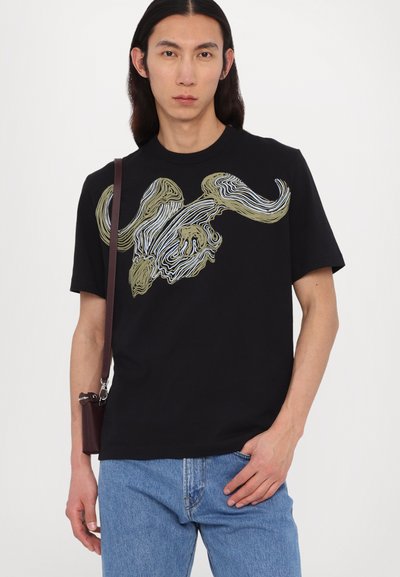 T-shirt noir avec un design graphique en or et des tourbillons blancs ressemblant à un taureau. Manches courtes, col rond, associé avec un jean bleu clair.