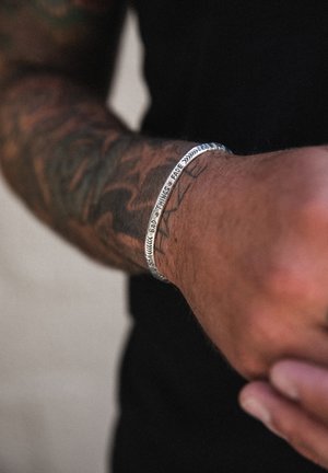 Bracciale d'argento con testo inciso, caratterizzato da una finitura liscia e una forma semicurva. Indossato su un polso tatuato.