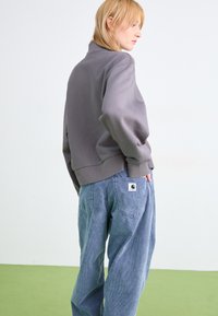 Hanorac gri cu guler înalt, cu o croială ușor supradimensionată, asortat cu o pereche de pantaloni lărgi din catifea cu aspect de bumbac, de culoare albastră, având buzunare la spate și un logo cusut.