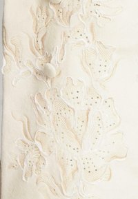 Textil de lana crema con intrincados bordados florales en beige suave y blanco, acentuado con pequeñas lentejuelas doradas. Detalle de superficie texturizada.
