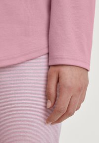 Langsleeve top van zachtroze stof met een gladde textuur, gecombineerd met roze-witte gestreepte onderstukken. Hand rustend aan de zijkant.