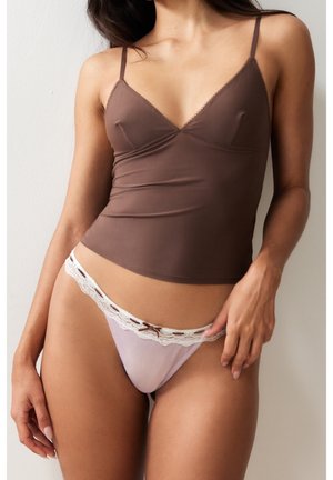 Femme portant un haut camisole marron et un string rose pâle orné de dentelle, debout contre un mur blanc uni.