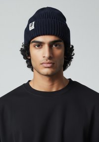 Laivastonsininen neulottu pipo, jossa on resorikuvio ja pieni brändilaatta, henkilö pukeutuneena mustaan paitaan, keskellä vaaleaa taustaa.