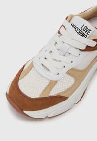 Sko med hvitt mesh-overdel, beige og brun semsket skinnakenter, runde lisser, logo på tungen, og en teksturert gummisåle.
