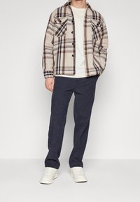 Kariertes Flanellhemd in Beige, Braun und Marineblau mit zwei Brusttaschen; kombiniert mit dunkelblauen Hosen und weißen Sneakers.