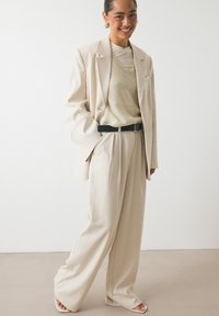 Costume beige avec un blazer ajusté et un pantalon ample. Associé à un pull beige clair et une ceinture noire, avec des textures lisses et douces.