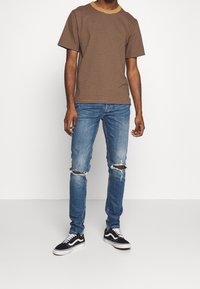 Hombre con camiseta a rayas marrón, jeans azules rasgados en las rodillas y zapatillas negras y blancas, de pie contra un fondo blanco.