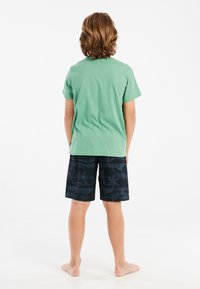 Groene katoenen T-shirt met korte mouwen, gecombineerd met zwarte shorts met een palmboompatroon, van achter bekeken. Blote voeten.