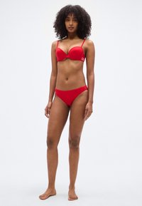 Κόκκινο σετ μπικίνι που περιλαμβάνει σουτιέν με ενίσχυση και ρυθμιζόμενες τιράντες και ταιριαστά μαγιό slip χαμηλοκάβαλα, και τα δύο από λείο, ελαστικό ύφασμα.