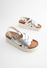 Hõbedased metallik sandaalid ristuva rihmaga, reguleeritava klambriga ja tekstureeritud korkplatoo tallaga. Omab kerget ja pehmet tallaosa.