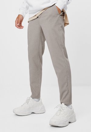 Mann trägt schlank geschnittene beige karierte Hose, weiße Socken und klobige weiße Sneaker, mit einer Hand in der Tasche.