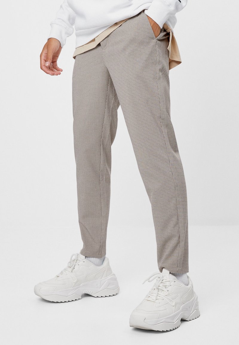 Homme portant un pantalon beige à carreaux slim, des chaussettes blanches et des baskets blanches épaisses, avec une main dans la poche.