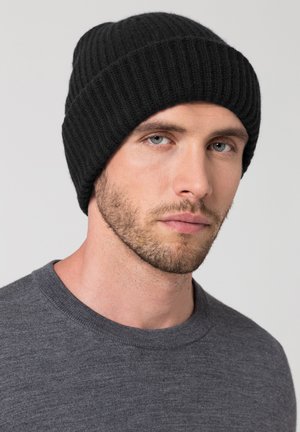 Schwarze gerippte Strickmütze mit einer weichen Textur. Eng am Kopf getragen, mit umgeschlagenem Schirm. Das Model trägt ein graues Crewneck-Shirt.