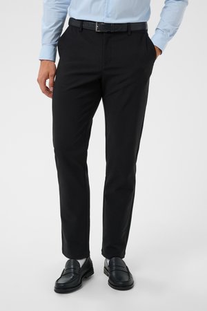 Hombre con pantalones de vestir negros, cinturón negro, camisa azul claro, calcetines blancos y mocasines negros, de pie con una mano en el bolsillo.