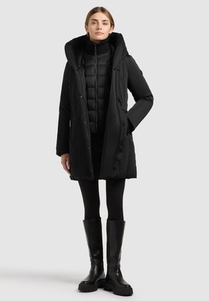 Joven mujer está de pie con un abrigo de invierno negro acolchado con capucha, leggings negros y botas negras hasta la rodilla sobre un fondo claro y sencillo.
