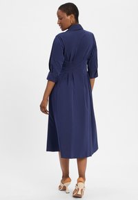 Granatowa sukienka midi z kołnierzykiem, rękawami 3/4, dopasowanym talią i plisowanym detalem z tyłu. Noszona z jasnymi sandałami na obcasie.