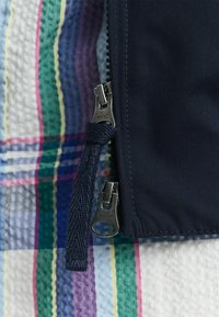 Tessuto blu navy con una superficie liscia, caratterizzato da una zip argentata e un tirazip intrecciato, posizionato su uno sfondo a quadri multicolore.
