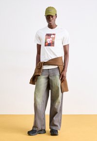 Άσπρο μπλουζάκι με φωτογραφική εκτύπωση, καφέ φούτερ δεμένο στη μέση, πράσινα φθαρμένα παντελόνια wide-leg, και μαύρα αθλητικά παπούτσια. Ελιά καπέλο.