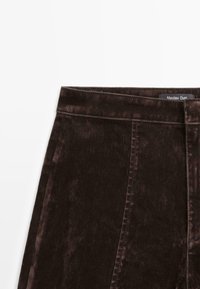 Pantaloni in denim marrone con una consistenza liscia, dotati di vita alta, leggero sbiadimento e dettagli di cucitura, contrassegnati da un'etichetta.