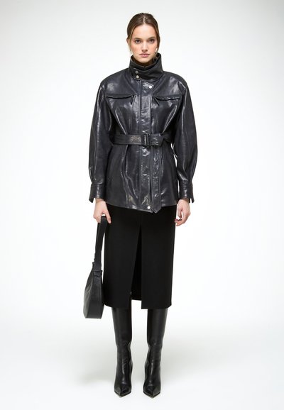 Vrouw draagt een zwarte leren jas met ceintuur, een zwarte midi-rok met een voorsplit, kniehoge zwarte laarzen en houdt een zwarte handtas vast.