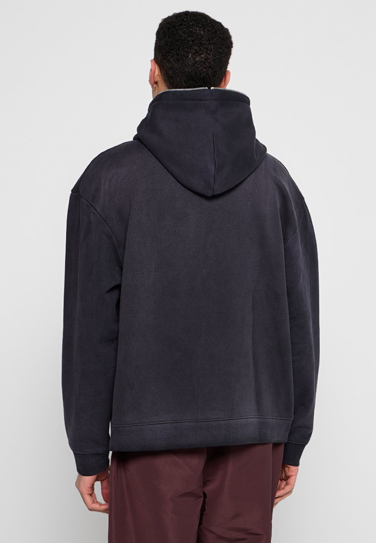 Dunkelgrauer Hoodie mit großer Kapuze, geripptem Material, überschnittenen Schultern und lockerer Passform, kombiniert mit kastanienbraunen Hosen.