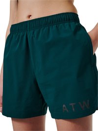 Zbliżenie osoby noszącej ciemnozielone szorty z elastycznym pasem i napisem „ATW” na lewej nodze, z ręką częściowo w kieszeni.