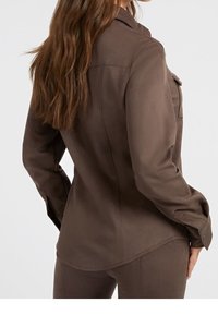 Chemise marron à manches longues avec col, poignets boutonnés et deux poches poitrine. Tissu doux au toucher lisse et coupe ajustée.