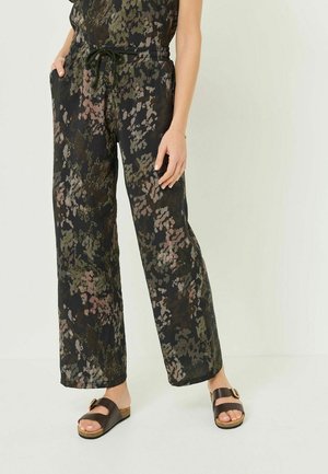 Kobieta ubrana w luźne spodnie w kwiatowy wzór z elastycznym paskiem z troczkiem w talii oraz ciemnobrązowe sandały z klamrą.