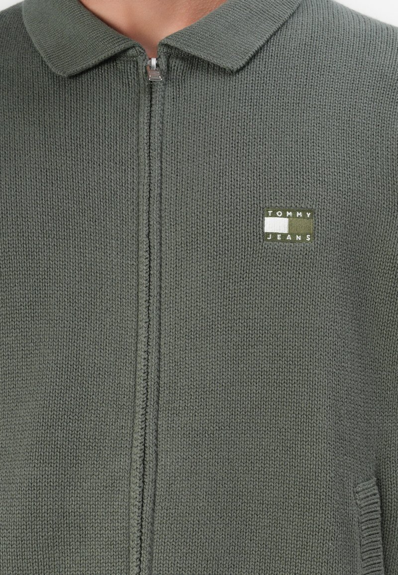 Green knitted cardigan with a zip closure and a collar. Features a small logo patch in the upper corner. Soft texture and casual design.