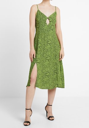 Femme portant une robe midi verte imprimé serpent avec bretelles spaghetti, fente latérale, découpe en forme de trou de serrure sur le devant et sandales noires à talons hauts.
