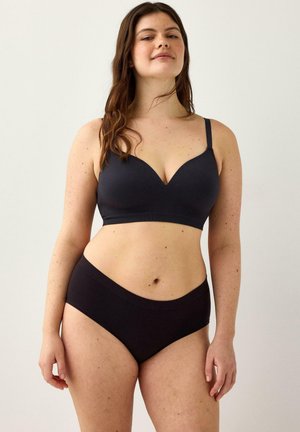 Donna che indossa un reggiseno nero e mutande abbinate, in piedi davanti a uno sfondo chiaro e uniforme con un'espressione sicura.